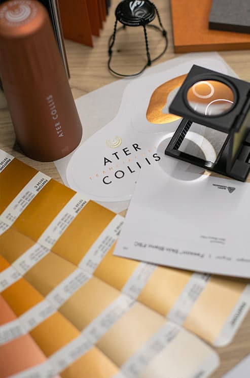 Primo piano di vari strumenti e materiali di progettazione, tra cui campioni di colore in tonalità oro, una lente di ingrandimento, una tazza metallica e carte stampate su una superficie di legno.