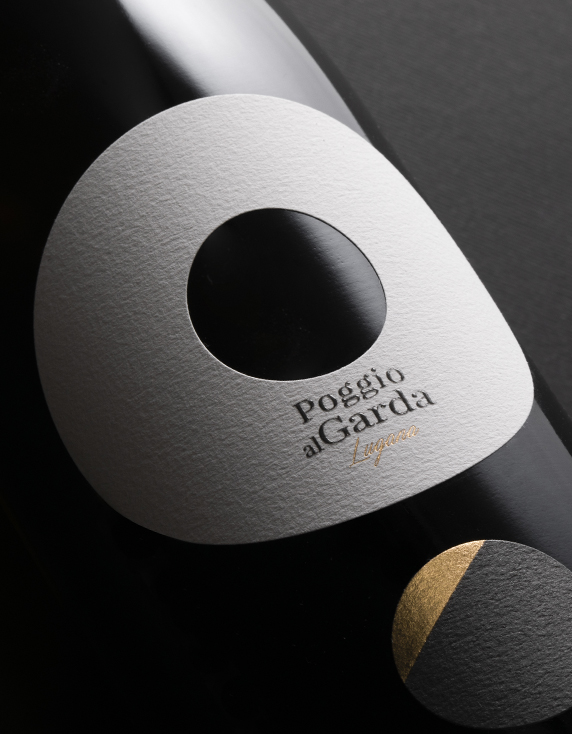 Primo piano di una bottiglia di vino etichettata Poggio al Garda Lugana con un moderno e minimalista disegno circolare bianco e oro su sfondo scuro.
